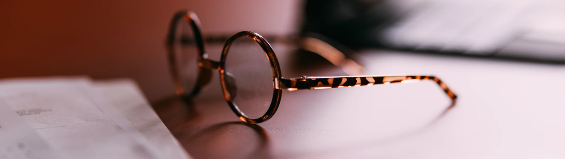 A pair of round, tortoise-shell eyeglasses rests on a desk next to a sheet of paper, with a soft, warm light casting a shallow depth of field.